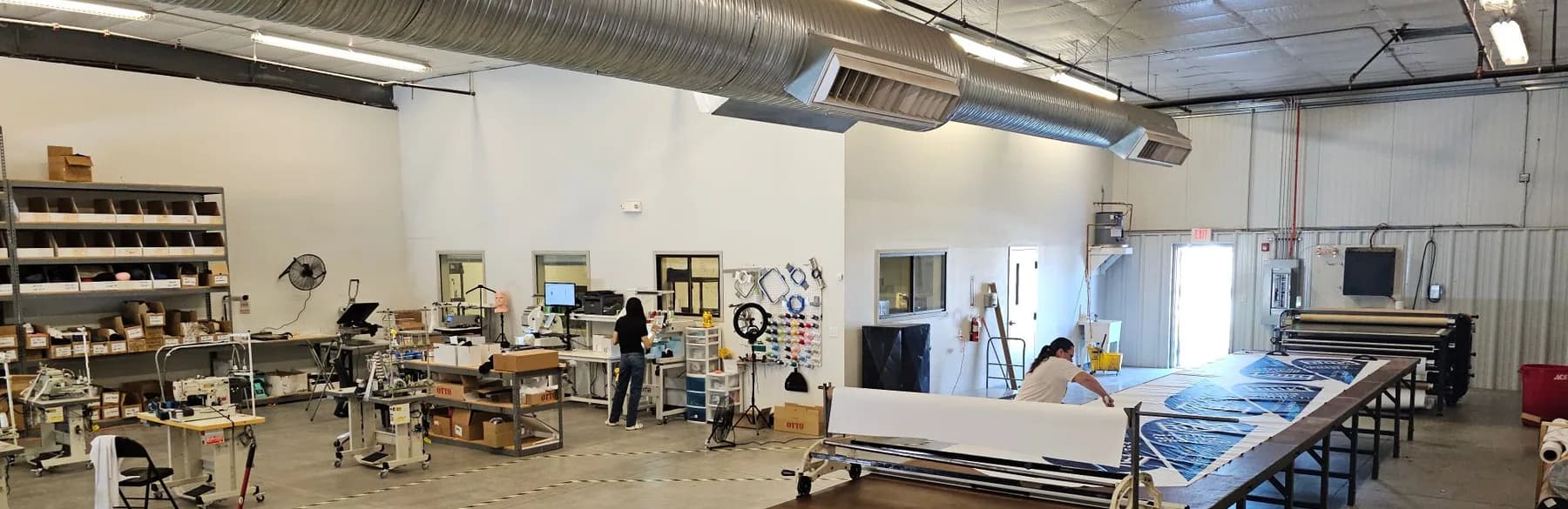 Our full shop floor: sewing stations, an embroidery and thread wall, a long cutting table, and a heat press under the ductwork